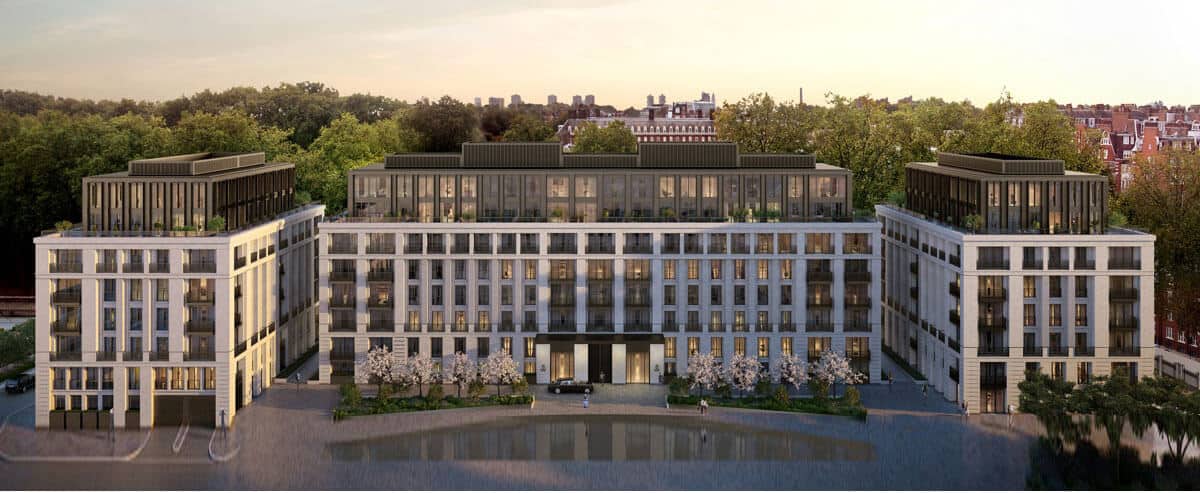 A modern, multi-story building with large windows and balconies, surrounded by greenery and trees, viewed from above at sunset.