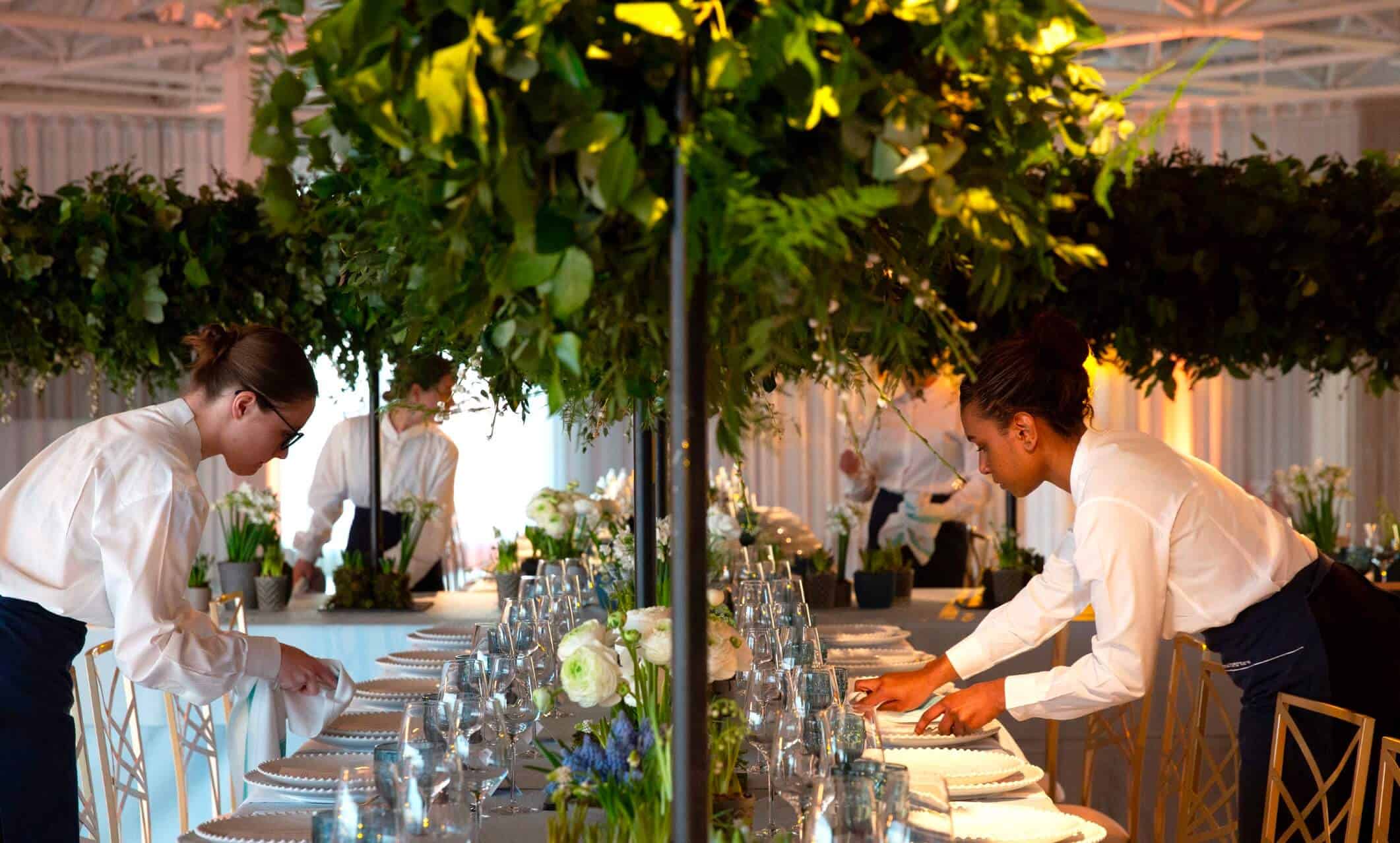 Two servers in white shirts are setting a long, elegant dining table decorated with greenery and floral arrangements, preparing for a formal event in a well-lit room.